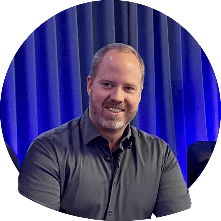 Andreas smilende i mørk skjorte, halvblondt hår og kort skjegg. Foto.